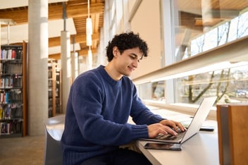 A young man with curly hair smiling as he uses a laptop at a library table, with bookshelves and large windows in the background.
