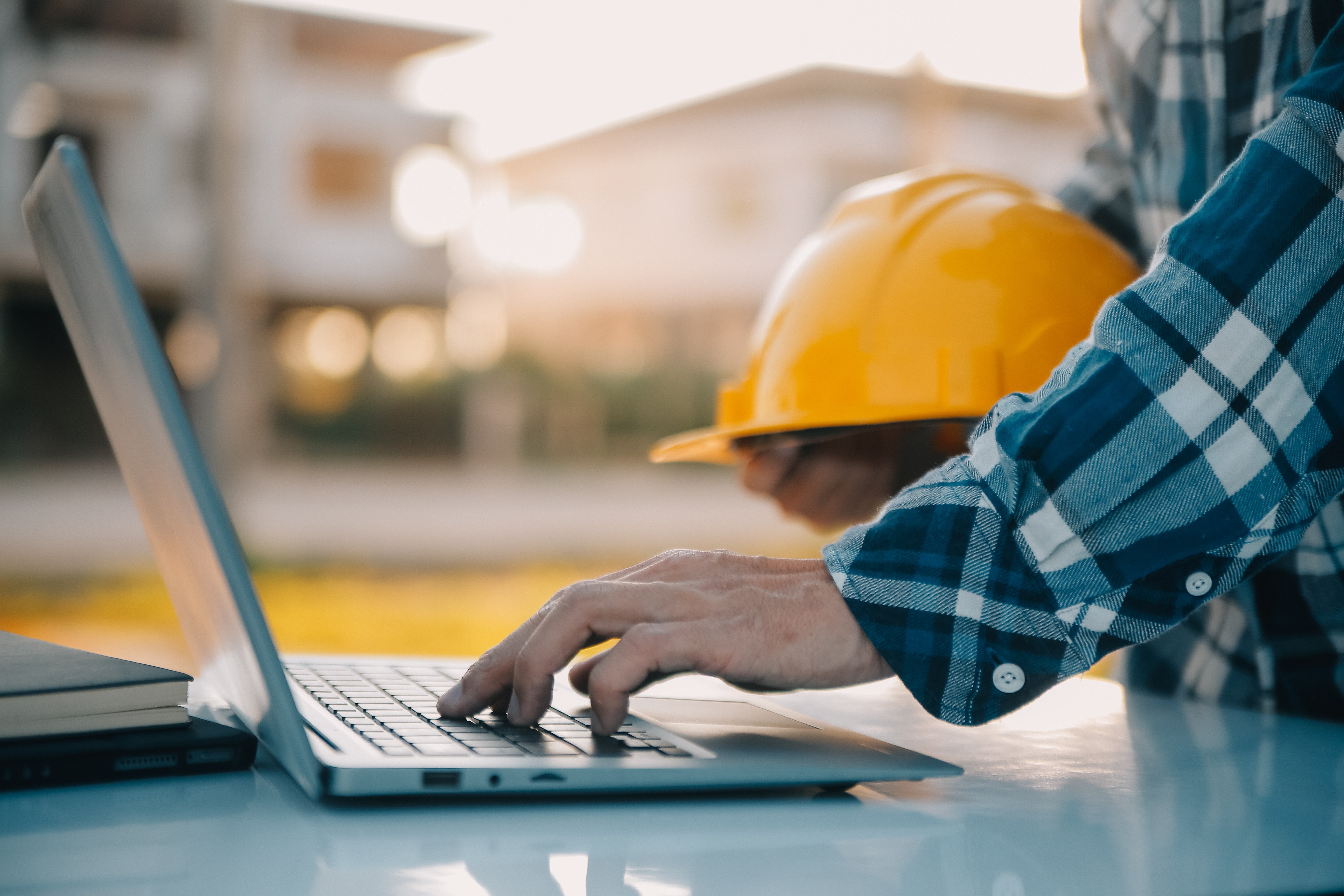 Person wearing a plaid shirt types on a laptop while holding a yellow hard hat. Blurry outdoor background.
